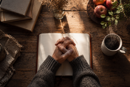 Hands clasped together resting on an open book at a kitchen table, early morning light