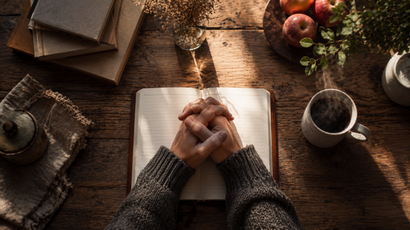 Hands clasped together resting on an open book at a kitchen table, early morning light