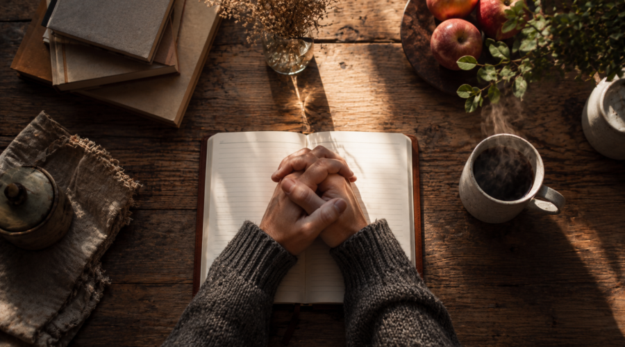 Hands clasped together resting on an open book at a kitchen table, early morning light