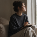 A person looking thoughtfully out a window with a journal resting on their lap
